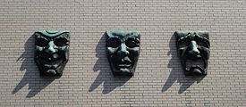 Drei Masken aus Bronze an einer weiß gestrichenen Backsteinwand. 