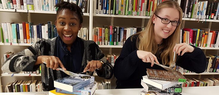 Zwei Mädchen mit Besteck in der Hand tun so, als würden sie einen Stapel Bücher essen. 
