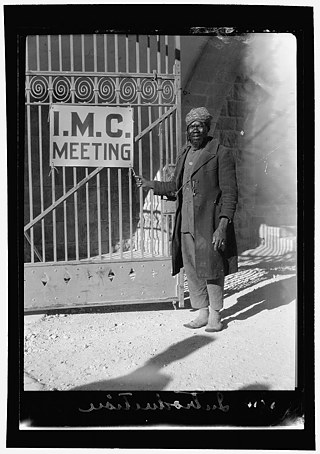 صورة بالأبيض والأسود لرجل يقف على يمين أحد المداخل يحمل لافتة كتب عليها "اجتماع المؤتمر التبشيري الدولي" سنة ١٩٢٨في فلسطين.