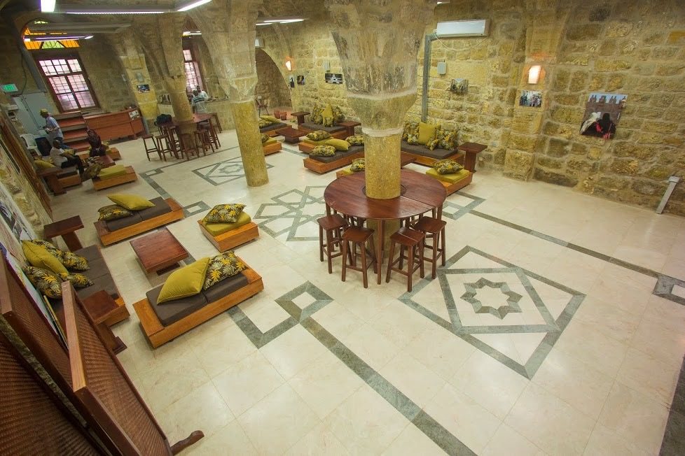 Image of the community center in Jerusalem in 2013. Stone floor, numerous tables and cushions on the floor to sit on, several columns in the room.