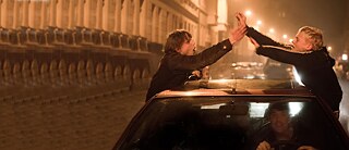 Scène du film 'Le temps des rêves'. De nuit dans une rue. Deux jeunes garçons debout, collés de chaque côté d'une voiture, leurs bras se rejoignent au dessus du toit de la voiture, un troisième assis au volant. Ils sont joyeux.