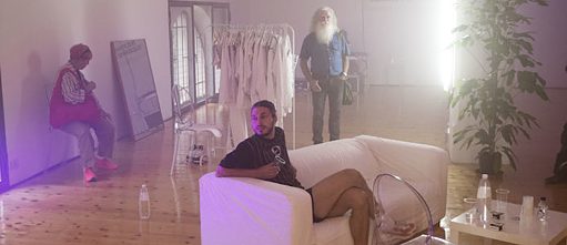Ausschnitt aus der Performance Purpose Next – Mann sitzt auf einem weißen Sofa, zwei weitere Menschen befinden sich stehend in dem Raum.