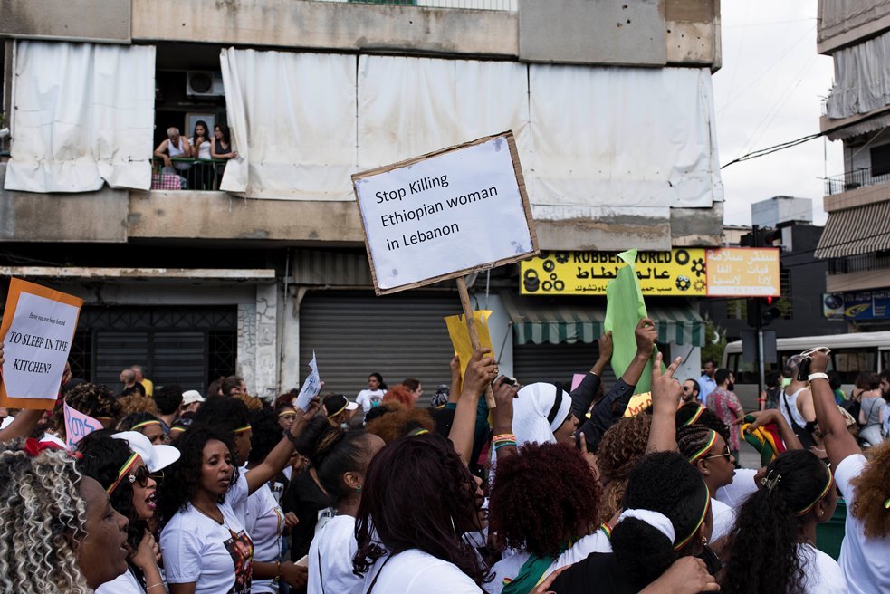 محتجون في بيروت، يرفعون لافتات من بينها "توقفوا عن قتل النساء الأثيوبيات في لبنان"، بعض الأشخاص يراقبون المنظر من النافذة.