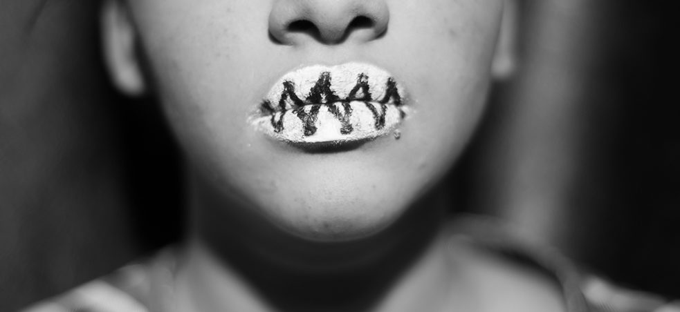 Photo en gros plan en noir et blanc d'un visage, lèvres peintes en blanc avec de la peinture noire entrecroisée, donnant l'impression que les lèvres sont cousues.