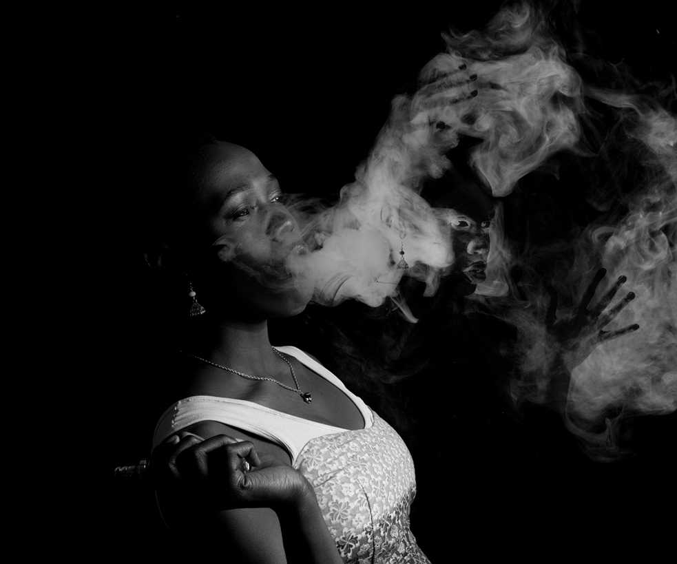 Black and white photo of woman looking into the distance, smoking, an image of her visible in the smoke.