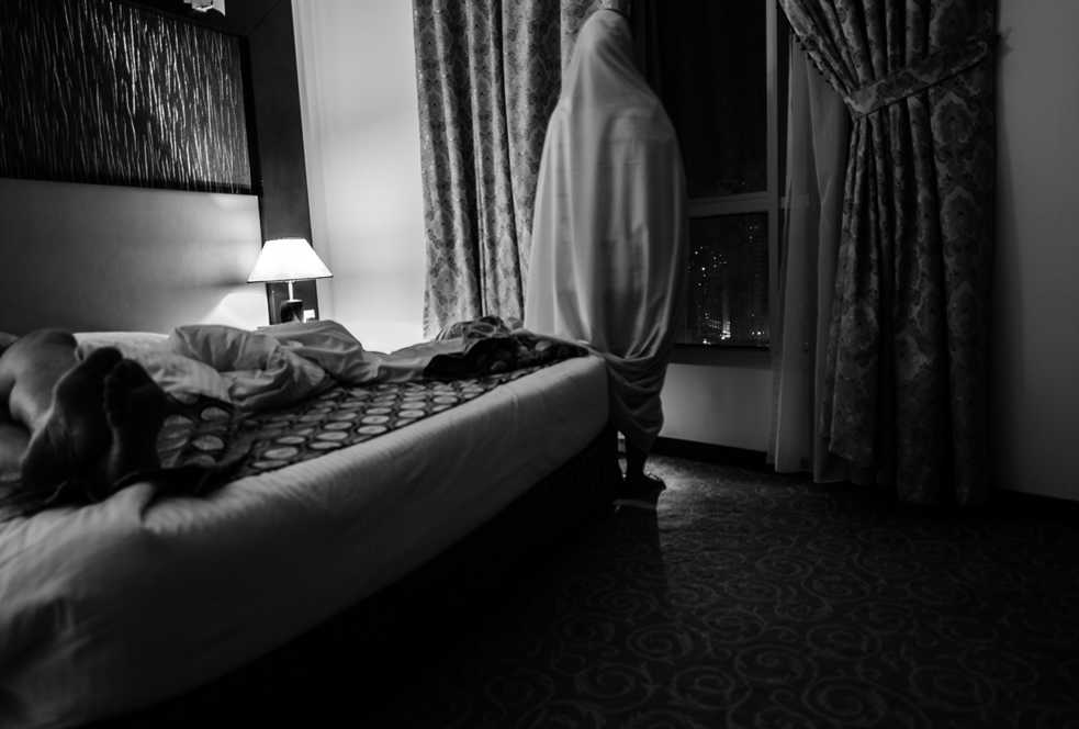 Black and white photo of a room, a covered women standing by the window looking out, a man lying on the bed.