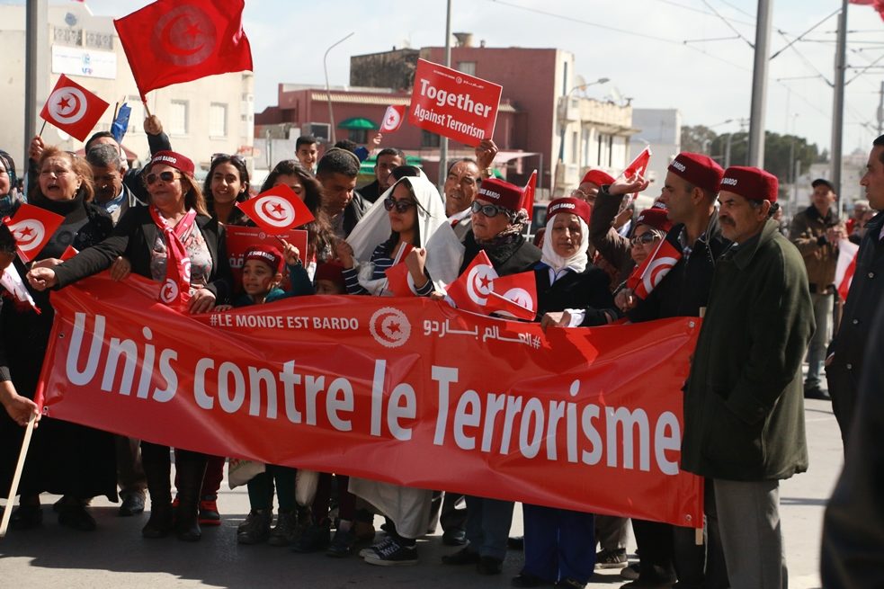 Un groupe de personnes, certaines portant la chéchia, le traditionnel chapeau de feutre rouge, brandissant une banderole qui dit  «Unis contre le terrorisme»