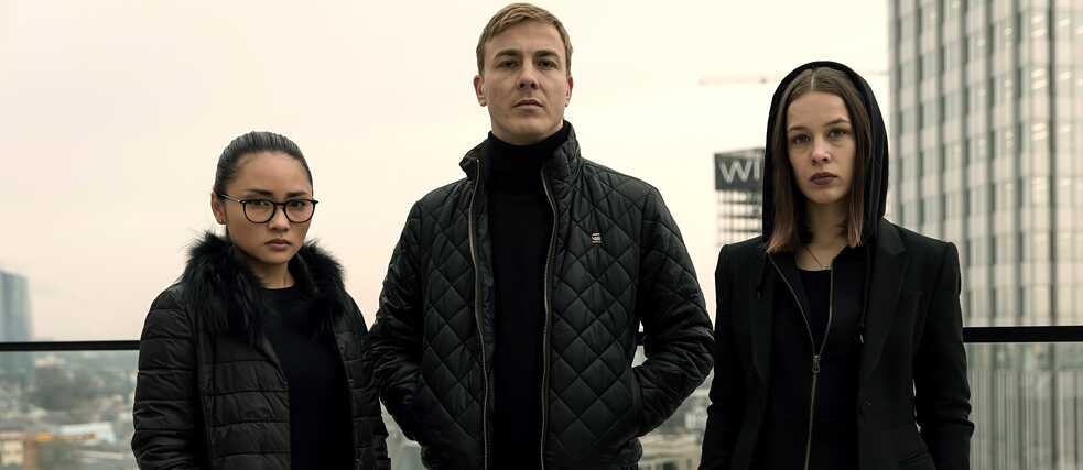 „Bad Banks - Die Höhle des Löwen“: Jana (Paula Beer), Adam (Albrecht Schuch) und Thao (Mai Duong Kieu) stehen, in Schwarz gekleidet, nebeneinander auf der Dachterrasse eines Hochhauses mit Blick auf Frankfurt und posieren für die Kamera.