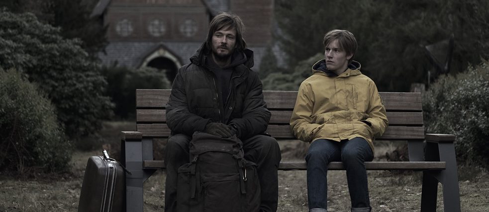 Un adolescent portant un imperméable et un homme barbu vêtu de noir sont assises sur un banc.