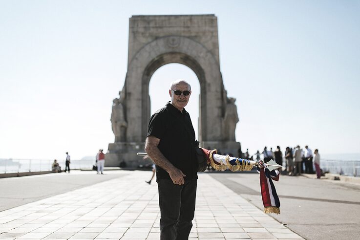 The pensioner Francis is standing in front of Porte d'Orient, holding a rolled-up flag and looking into the camera.