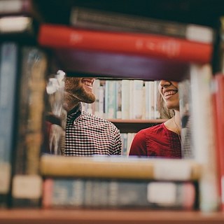 Door een spleet in een boekenkast lachen een mannengezicht en een vrouwengezicht elkaar toe. 