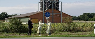 Zwei junge Frauen in Schutzkleidung vor einem Gebäude auf einem Feld, neben ihnen ein Mann