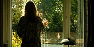 Still frame from the ZDF Series "Perfume" - "Perfume - Ambra": Elena Seliger (Natalia Belitski) looks through the patio door into the garden, where the little neighbor boy stands, who returns her gaze and holds something orange in her hand.