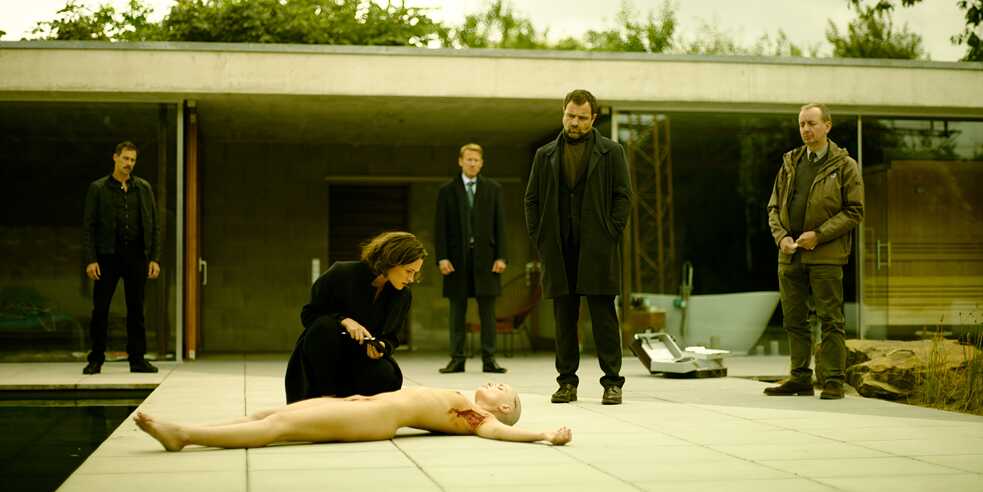 "Parfum - Ambra": Brettschneider (Marc Hosemann), Grünberg (Wotan Wilke Möhring) y Köhler (Juergen Maurer) están de pie en la terraza de un bungalow. Allí, un cadáver femenino desnudo con cráneos esquilados yace junto a un estanque. Nadja Simon (Friederike Becht) se arrodilla delante de ella y la mira de cerca.
