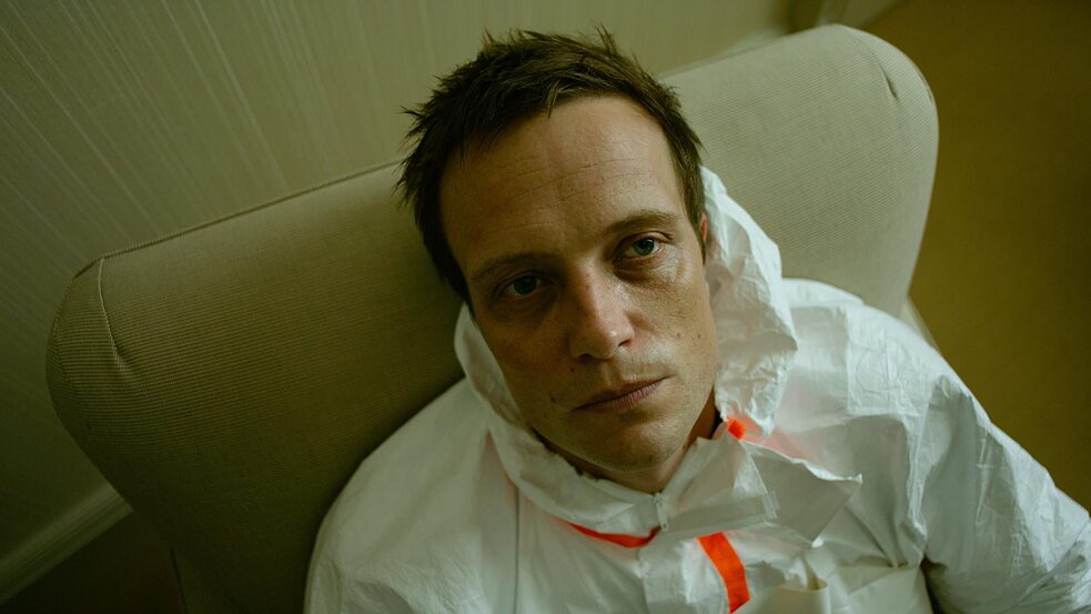 "Parfum - Die dritte Substanz": Close-up de Moritz de Vries (August Diehl) sentado en un sillón con un traje protector blanco y mirando fijamente a la cámara con una mirada vacía.