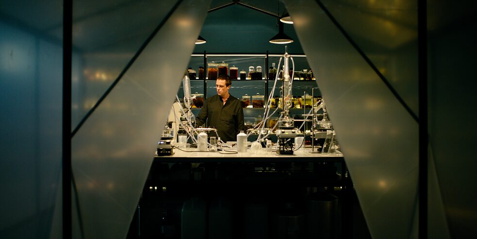Still frame from the ZDF Series "Perfume" - "Perfume - Ambra": Moritz de Vries (August Diehl) is standing in his laboratory with equipment for extraction on the table in front of him. Behind him stands a shelf, with different glasses of unknown content.