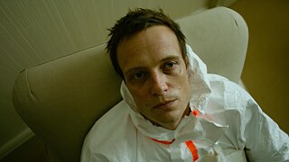 Still frame from the ZDF Series "Perfume" - "Perfume - The Third Substance": Close-up of Moritz de Vries (August Diehl) sitting in a white protective suit on a chair and staring blankly at the camera.