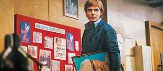 Ein Mann steht vor einer Wand mit Plakaten aus der DDR und hält ein großes Porträt eines Mannes unter seinem Arm.