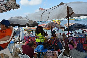 A crowded beach, and a women in a bright yellow vest talking to people.