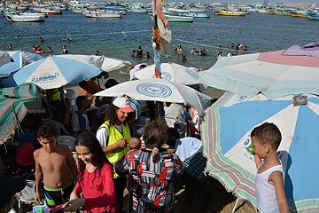 A crowded beach, and a women in a bright yellow vest talking to a women and her child.