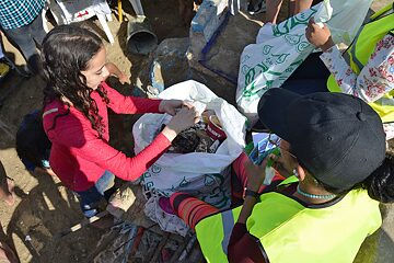 Multiple people around a plastic bag filled with rubbish, a young girl putting more rubbish in the bag.