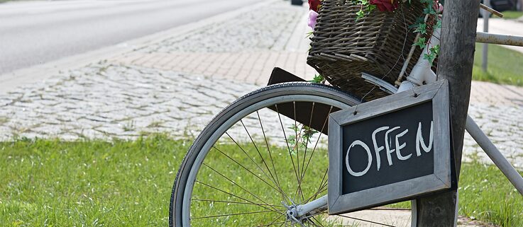 Schild mit Aufschrift „offen” am Lenker eines an einen Pfosten gelehnten Fahrrades