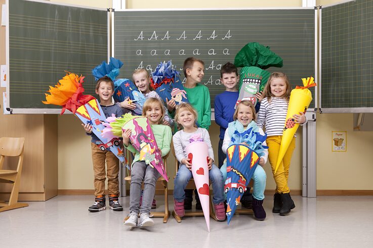 Mehrere Erstklässler stehen am ersten Schultag mit ihren Zuckertüten im Arm vor der Tafel im Klassenraum. Sie lachen und sind fröhlich.