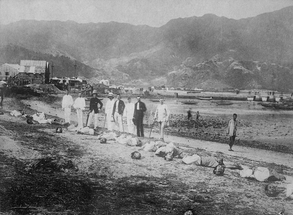 Kiauchau, antiga colônia alemã na China: chineses decapitados (supostamente criminosos) à beira da estrada. Atrás deles, um grupo de europeus posa para uma foto em grupo. Fotografia tirada por volta de 1900.