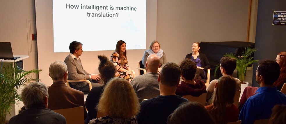 Vier Referent*innen mit Publikum, an der Wand die Frage "How intelligent is machine translation?"