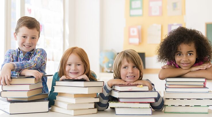 Vier Kinder sitzen in einem Klassenraum vor Bücherstapeln und lächeln. Sie stützen sich mit den Armen auf den Büchern ab. 