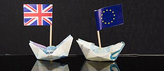 Zwei aus Eurobanknoten gefaltete Schiffchen, eins mit Europaflagge, eins mit UK-Flagge, vor schwarzem Hintergrund 