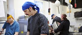 Mehrere Köche arbeiten in einer Restaurantküche. Sie tragen Kopftücher und Kochjacken.