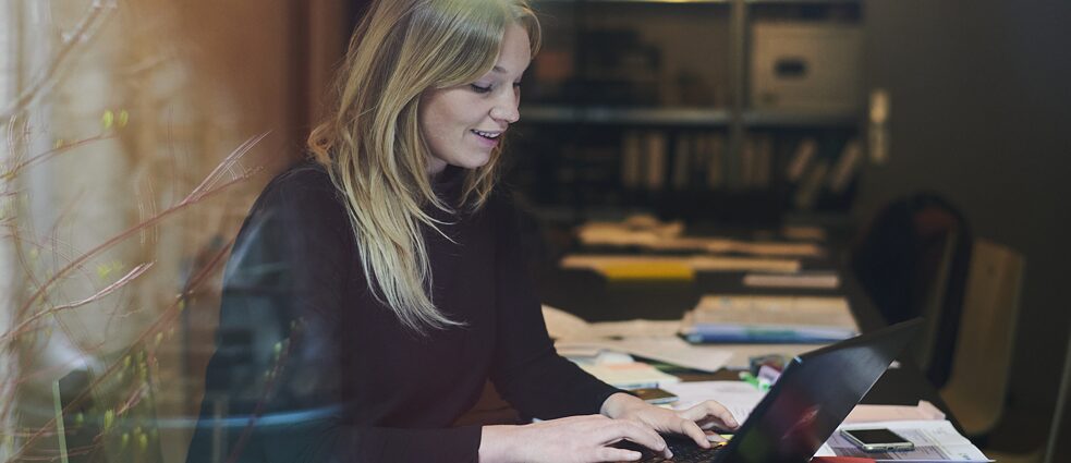 Eine Frau sitzt an einem Tisch in ihrer Wohnung und arbeitet am Laptop.
