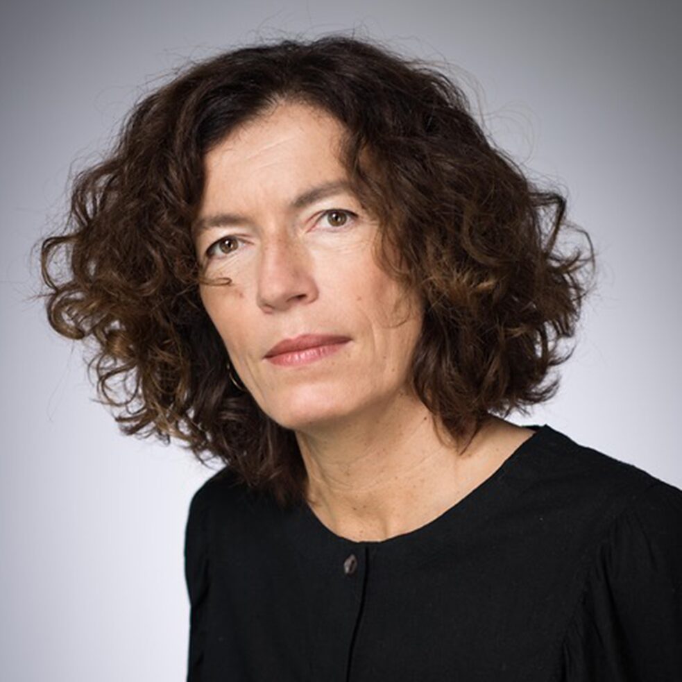 Portraitbild von Anne Weber vor grauem Hintergrund; sie hat lockige braune Haare und trägt einen schwarzen Pullover