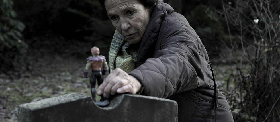 DARK Eine alte Frau stellt eine Spielzeugfigur auf einen Grabstein
