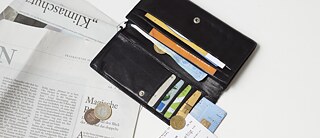 Coins, banknotes and credit cards can be seen in an open purse. The purse is lying on a newspaper.