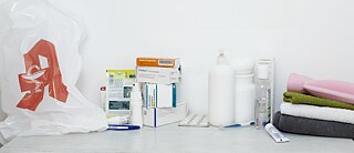 On a table is a bag with the pharmacy symbol. Next to it are various medicines, towels and a hot water bottle.