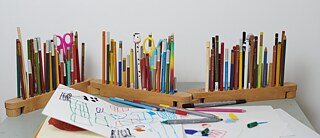 On a table are pencil holders with many colourful pens and craft scissors. Next to them are self-painted pictures by children.