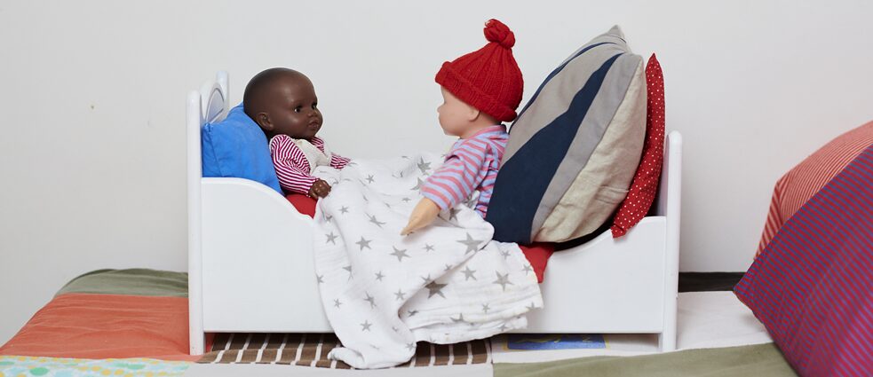 zwei Puppen mit unterschiedlicher Hautfarbe sitzen sich in einem Puppenbett gegenüber.
