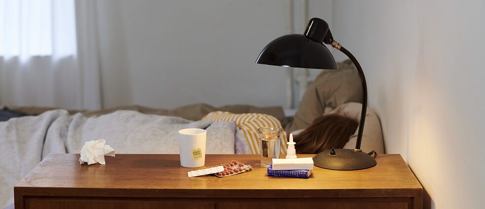 Eine Person liegt krank im Bett. Auf dem Nachttisch sind Taschentücher, Nasenspray, Medikamente und eine Tasse Tee zu sehen.