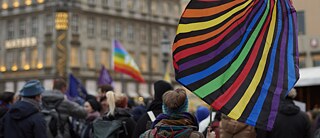 A demonstration with many people can be seen. A rainbow flag can be seen in the foreground.