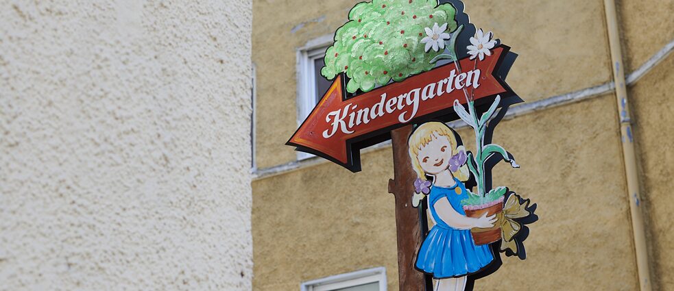Ein Schild, auf dem Kindergarten geschrieben steht, führt ein Pfeil mit einem gezeichneten Mädchen in Richtung des Kindergartens.