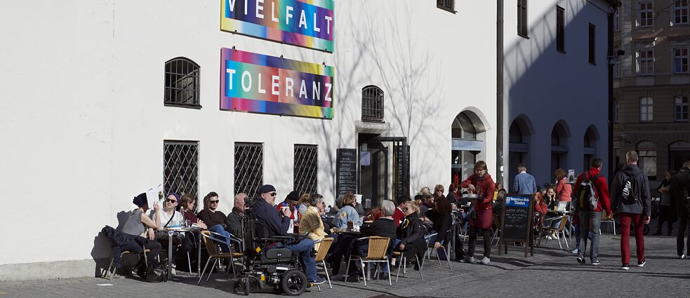 Menschen sitzen vor einem Cafe in der Sonne. Ein Mann in einem Rollstuhl ist zu sehen.