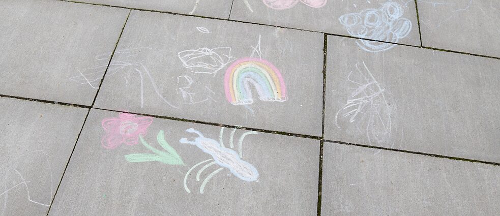 Auf einer Straße sind mit Straßenkreide eine Blume, ein Regebogen und eine Spinne gemalt.