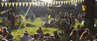 Ansicht auf eine sommerliche Party auf einer Wiese mit vielen Menschen, Essensständen und Dekoration