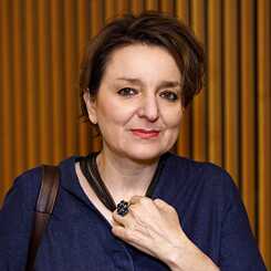 Quadratisches Bild von Eva Illouz vor bräunlichem Hintergrund; sie hat schwarze, kurze Haare und hält ein Glas in der linken Hand