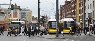 You see a street in Berlin with many people walking across the street and a bus and trams in the background.