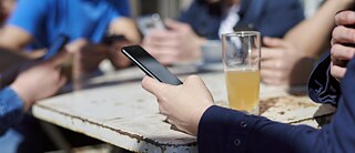 Close-up on an outdoor table with a drink on it. One arm of a person operating a smartphone is seen.
