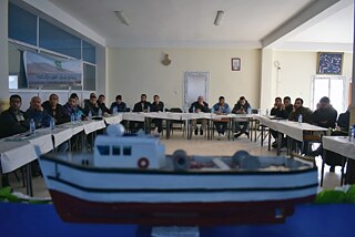 Conference room with people sitting around tables, a miniature boat in the foreground.