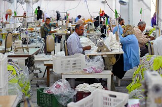 Trabalhadoras e trabalhadores na oficina de costura da empresa egípcia Sekem, onde costuram pequenos produtos têxteis e componentes para bonecas de pano, fotografados em 27/10/2014. A iniciativa Sekem é uma empresa ligada à antroposofia fundada em 1977 por Ibrahim Abouleish. A empresa desenvolveu uma área desértica localizada a cerca de 47 km a nordeste do Cairo para a agricultura biodinâmica e desde então tem sido capaz de se expandir continuamente em outras áreas. A companhia, que se tornou uma holding composta por seis grupos de empresas, opera comércio justo e mantém uma escola, um jardim de infância, um centro de treinamento profissional, uma clínica e uma universidade para seus quase 2 mil funcionários, funcionárias e suas famílias. Em 2003, Ibrahim Abouleish recebeu o Prêmio Nobel Alternativo pela iniciativa Sekem.